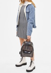 Svart halvgenomskinlig ryggsäck med ett handtag upptill och en ytterfick med dragkedja. Buren med en blå kordjacka och rutigt klänning. Vita snörboots.