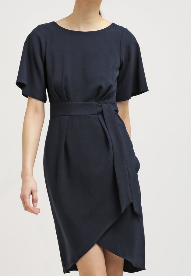 Femme portant une robe bleu marine foncé longueur genou avec manches courtes, ceinture à la taille et jupe portefeuille asymétrique sur un fond uni.
