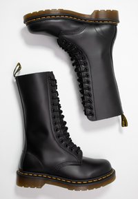 Botas de combate de cuero negro con múltiples ojales, suela de goma gruesa, costuras amarillas y una lengüeta para facilitar el calzado. Altura hasta la mitad de la pantorrilla.
