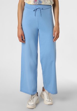 Donna che indossa pantaloni larghi color azzurro chiaro con coulisse in vita e sneakers bianche chunky, in piedi davanti a uno sfondo semplice.