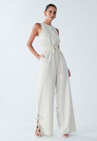 Brezrokavnik bež kombinezon z elastičnim pasom, stranskimi žepi in razporki, ki so zavezani z blago okoli gležnjev. Gladek, lahek material.