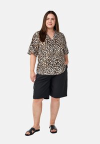 Kort-日本se luipaardprint shirt in beige en bruin, gecombineerd met zwarte shorts. Model draagt zwarte sandalen met decoratieve studs.
