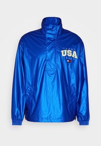 Lett blå overtrekksjakke med høy hals, elastiske mansjetter og sidelommer. Har "TOMMY USA"-logo i grønn og hvit. Vanntett materiale.