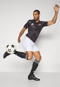 Maillot de football à rayures noires et rouges à manches courtes, shorts blancs, chaussures de football noires, chaussettes noires avec des rayures blanches, en train de frapper un ballon de football à motifs.