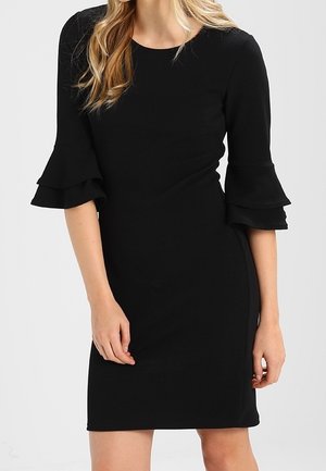 Kvinde iført en tætsiddende sort kjole med lagdelte klokkeærmer, stående med hænderne afslappet langs siden mod en neutral baggrund.