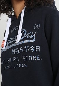 Persona que lleva una sudadera negra de Superdry con cordones blancos y texto en japonés y en inglés en el frente.