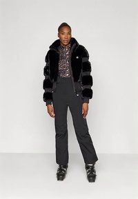 Γυναίκα φορώντας μαύρη ψεύτικη γούνα, μπλούζα με λεοπάρ σχέδιο, μαύρο παντελόνι σκι και μαύρες μπότες σκι, στέκεται μπροστά σε απλό ανοιχτό φόντο.