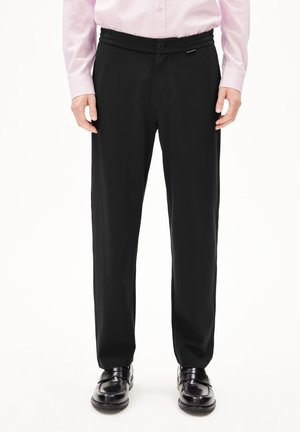 Schwarze Hose mit glatter Textur, geradem Schnitt, elastischem Bund und Knopfverschluss. Kombiniert mit polierten schwarzen Schuhen.