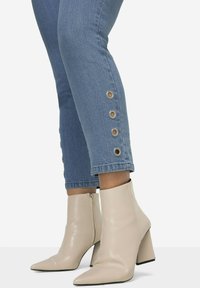Botas beige hasta el tobillo con puntera afilada y tacón grueso, combinadas con jeans de lavado claro que presentan detalles de ojetes de metal en el dobladillo.