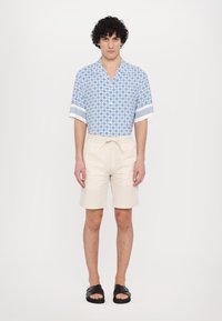 Cămașă cu mânecă scurtă, de culoare albastră, cu un model geomeric și tiv alb; pantaloni scurți crema, dintr-un material ușor; sandale negre.