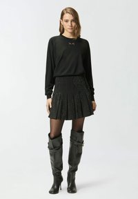 Top nero a maniche lunghe con un logo discreto, abbinato a una gonna nera plissettata con dettagli traforati e stivali neri in pelle alti fino al ginocchio.