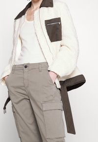 Krémová bunda z faux beránka se hnědou zipovou kapsou, spárovaná s olivovými cargo kalhotami s bočními kapsami a volným střihem.