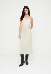 Midi φόρεμα με λαιμόκοψη cowl, χωρίς μανίκια, από ελαφρύ μπεζ ύφασμα, με ζώνη που δένει στη μέση και ίσια γραμμή, συνδυασμένο με μαύρες μπότες με τακούνι.