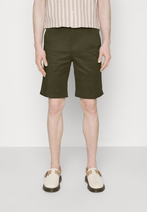 Personne portant un short vert foncé arrivant au genou, une chemise rayée boutonnée rentrée dans le pantalon, et des chaussures blanches à enfiler avec des boucles.