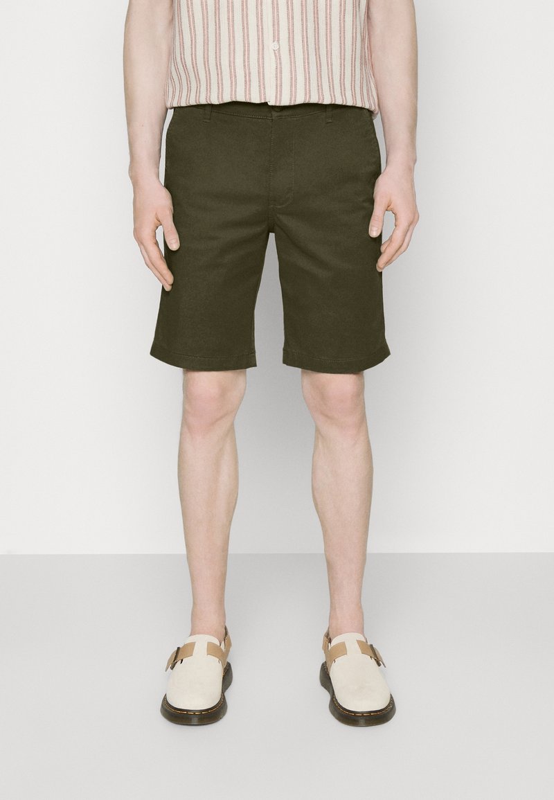 Personne portant un short vert foncé arrivant au genou, une chemise rayée boutonnée rentrée dans le pantalon, et des chaussures blanches à enfiler avec des boucles.