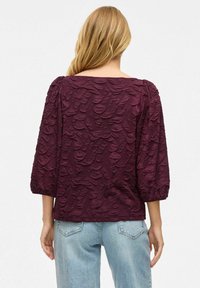 Bordová blůza s texturovanou látkou a vystouplými vzory. Navržena se širokým výstřihem a rukávy po tři čtvrtě, nošena s světle modrými džíny.