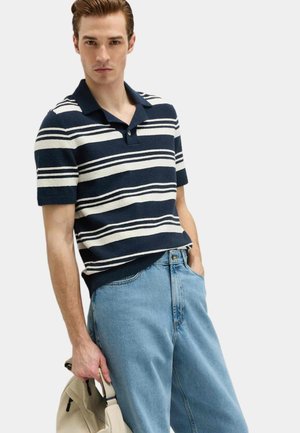 Giovane uomo che indossa una polo a maniche corte a righe blu navy e bianche e jeans azzurri, con una borsa beige in mano e l'altra mano in tasca.