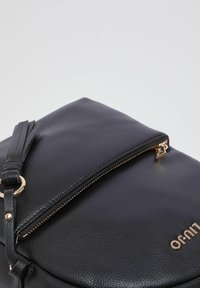 Sac en cuir noir avec une finition texturée, des fermetures éclair et des accents dorés. Il possède un rabat supérieur et une bandoulière amovible avec un détail à pampille.