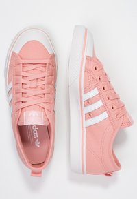 Un paio di sneakers Adidas in tela rosa con strisce bianche e suole in gomma, una scarpa mostrata dall'alto e l'altra di lato.