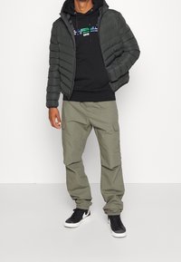 Chaqueta acolchada negra con capucha, combinada sobre una sudadera negra con texto de colores, acompañada de pantalones cargo oliva y zapatillas negras.