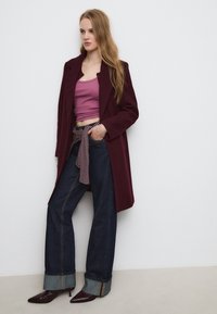 Burgunderfarbener langer Mantel über einem rosa verkürzten Tanktop, kombiniert mit dunklen, weiten Jeans, die mit einem gestreiften Gürtel und umgekrempelten Säumen versehen sind. Spitz zulaufende burgunderfarbene Absätze.