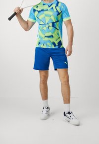 Camiseta deportiva de Puma con un patrón geométrico en azul y verde, combinada con pantalones cortos azules, zapatillas de tenis blancas y sosteniendo una raqueta.