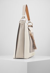 Krämfärgad handväska med texturerad läderfinish, med kontrasterande mörka detaljer, guldhardware och en dekorativ tofsdetalj.