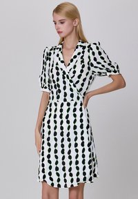 Robe portefeuille blanche avec un motif de pois noirs et verts, manches courtes bouffantes, et une taille cintrée avec une jupe évasée. Texture de tissu lisse.