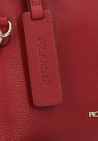 Borsa a tracolla in pelle rossa con texture, dotata di un'etichetta logo impressa con la scritta "PICARD SINCE 1928" e un accento in hardware dorato.