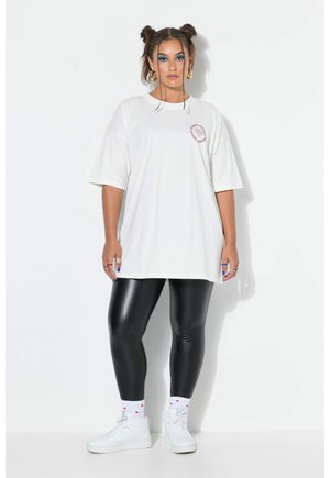 Frau mit zwei Dutts, die ein übergroßes weißes T-Shirt, schwarze glänzende Leggings, weiße High-Top-Sneaker, goldene Creolen und kräftiges blaues Augen-Make-up trägt.