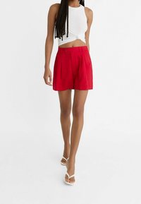 Shorts rouges taille haute avec des plis, fabriqués à partir d'un tissu léger. Associés à un débardeur blanc court et des sandales blanches.