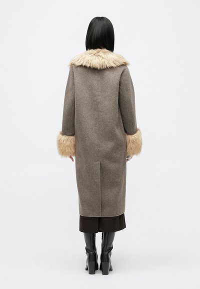 Μακρύ καφέ παλτό με ψεύτικη γούνα στον γιακά και τα μανίκια, μαλακή υφή, ίσια γραμμή, με άνοιγμα στο πίσω μέρος, φορεμένο πάνω από μια σκούρα φούστα και ψηλές μαύρες μπότες με τακούνι.