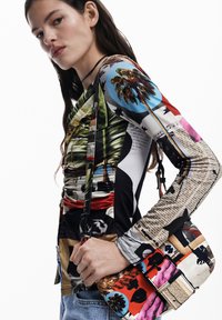 Mehrfarbige Langarmoberteil mit grafischen Drucken und Texturen; das Modell hält eine gemusterte Handtasche mit lebhaften Farben und Designs.