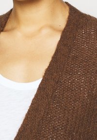 Hnědý pletený cardigan s texturovaným vzorem, s širokým otevřeným předním dílem a kontrastem s bílým tričkem.