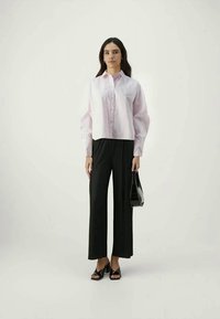 Light pink button-up shirt with a relaxed fit, paired with black pleated wide-leg trousers. Model holds a small black handbag and wears black sandals.