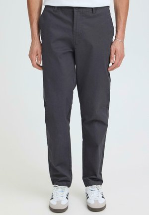 Mann trägt dunkelgraue, gerade geschnittene Hose mit Seitentaschen und weiße Turnschuhe, steht vor einem schlichten hellen Hintergrund.