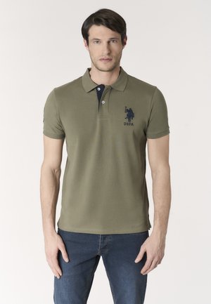 Uomo che indossa una polo USPA verde oliva e jeans blu, in piedi davanti a uno sfondo chiaro e uniforme.