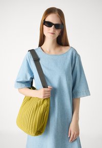 Sac plissé jaune avec une surface texturée, tenu par une personne portant une robe en denim bleu clair, dotée d'une sangle réglable grise.