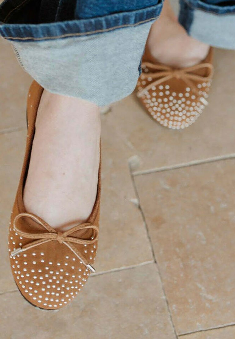 Bailarinas de ante marrón con lunares blancos, un lazo en la parte delantera y puntera redondeada. Llevadas con jeans de mezclilla remangados.