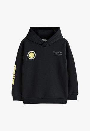 Ragazzo giovane con capelli ricci che indossa una felpa con cappuccio nera con una faccia sorridente gialla e pantaloni cargo scuri, in piedi davanti a uno sfondo chiaro e semplice.