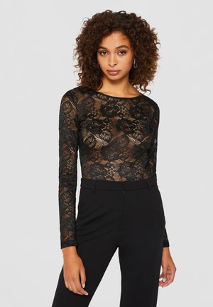 Žena s kovrčavom kosom u crnoj cvjetnoj čipkastoj majici dugih rukava i crnim hlačama visokog struka, stoji na jednostavnoj svijetloj pozadini.