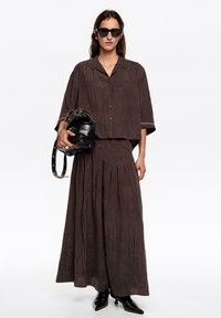Brązowy lekki, teksturowany komplet dwuczęściowy z krótkim rękawem i zapinanym na guziki topem oraz długą plisowaną spódnicą; dopełniony czarną torebką i okularami przeciwsłonecznymi.