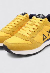 Scarpe da ginnastica gialle con materiale in suede e rete, dotate di punta rotonda, design con lacci, dettagli in blu navy e logo geometrico sul lato.