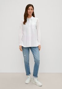 White long-sleeve shirt with a chest pocket, paired with light blue distressed jeans and white sneakers with green accents.