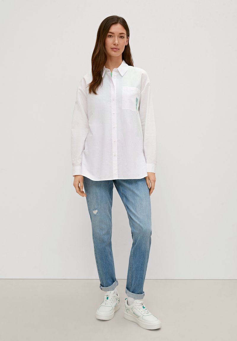White long-sleeve shirt with a chest pocket, paired with light blue distressed jeans and white sneakers with green accents.