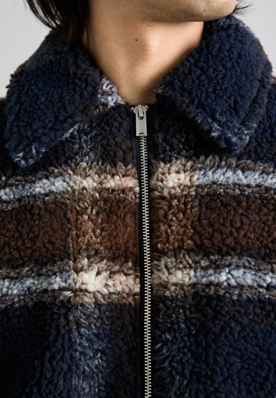 Casaco de fleece com um padrão de xadrez em azul marinho, castanho e creme. Apresenta fecho de zíper e gola. Tecido suave e texturizado.