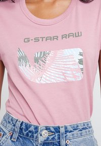 Růžové tričko s krátkým rukávem vyrobené z bavlny, s grafickým designem G-Star Raw a pastelovými palmami na přední straně. Viditelné džíny šortky.