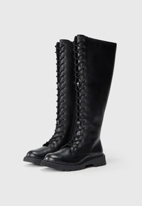 Alte scarpe in pelle nera con lacci e suole spesse, con zip laterali, in posizione eretta su uno sfondo chiaro.