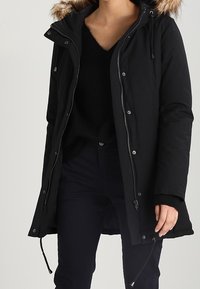Zwarte, knielange parka jas met een gevoerde capuchon van bont, ritsluiting, trekkoorden en zijzakken; gemaakt van een gladde, waterafstotende stof.