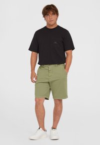 Schwarzes Kurzarm-T-Shirt mit Tasche, kombiniert mit olivgrünen Shorts. Das Modell trägt weiße Sneakers mit schwarzen Akzenten. Neutraler Hintergrund.